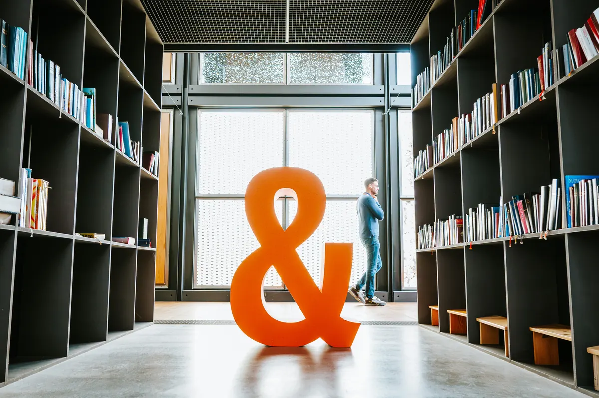Ein großes orangefarbenes Und-Zeichen steht zwischen hohen Bücherregalen